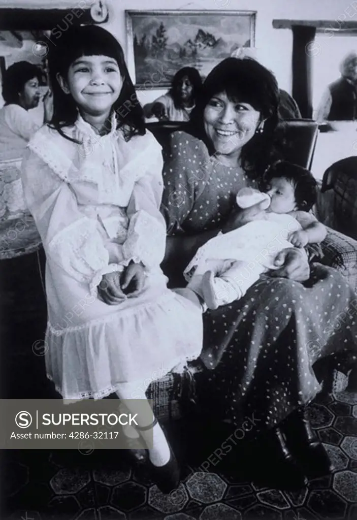 Young Native American Pueblo girl in a white dress smiling and sitting next to her mother as she feeds her young baby with a bottle.