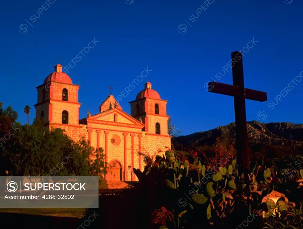 Sun shining on Mission Santa Barbara at sunrise, with cross in foreground.