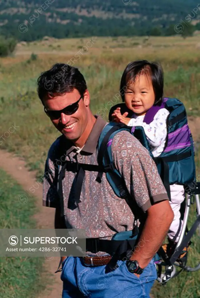 Father carrying adopted young Asian American daughter on his back in backpack.
