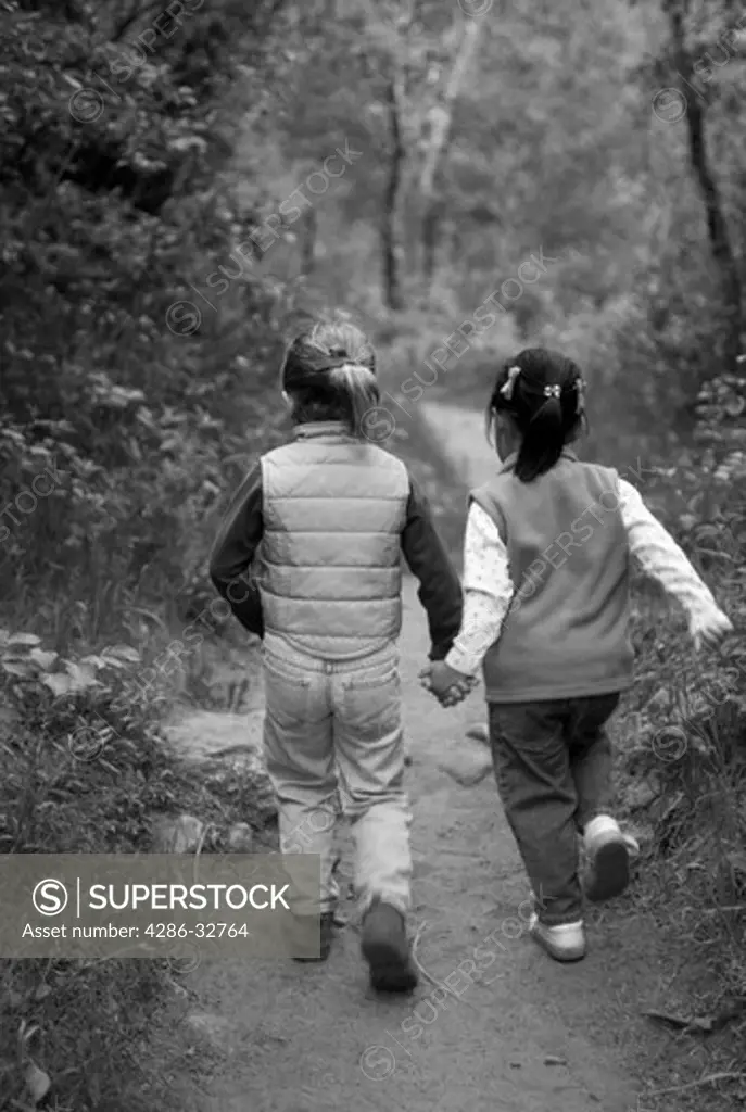 Black and white image of two young girls holding hands and hiking on a path through the forest.