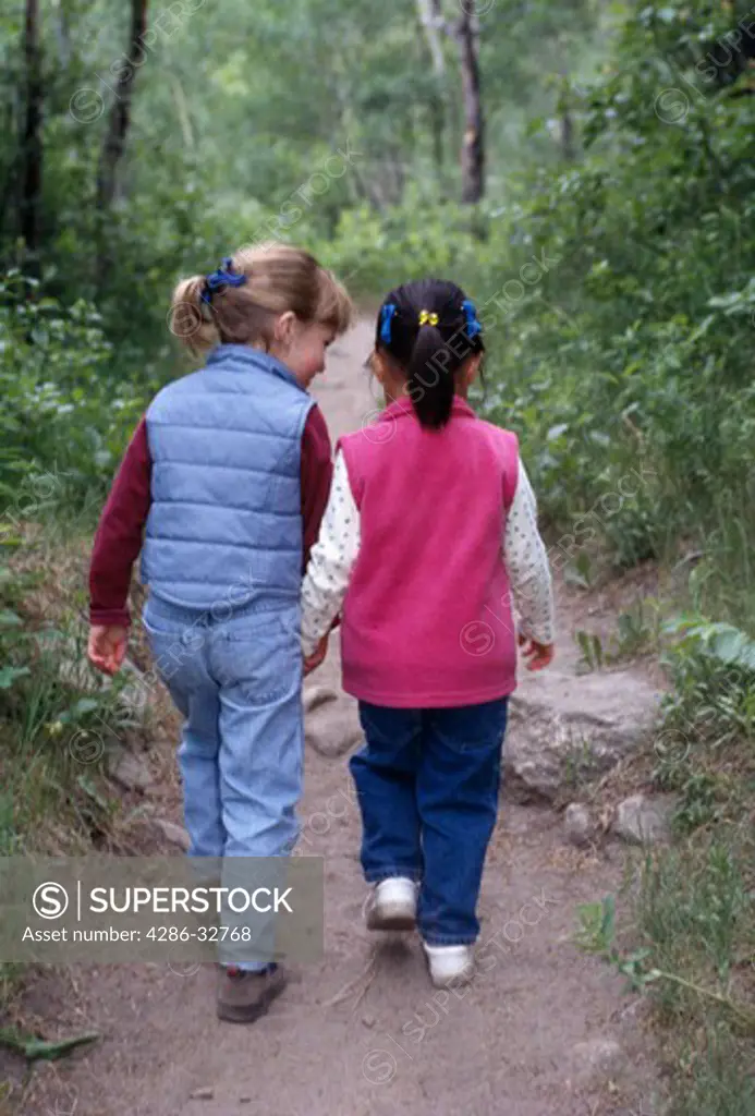 Two young girls holding hands and hiking on a path through the forest.