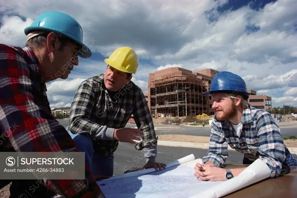 Two construction workers and their supervisor discuss plans for office building.