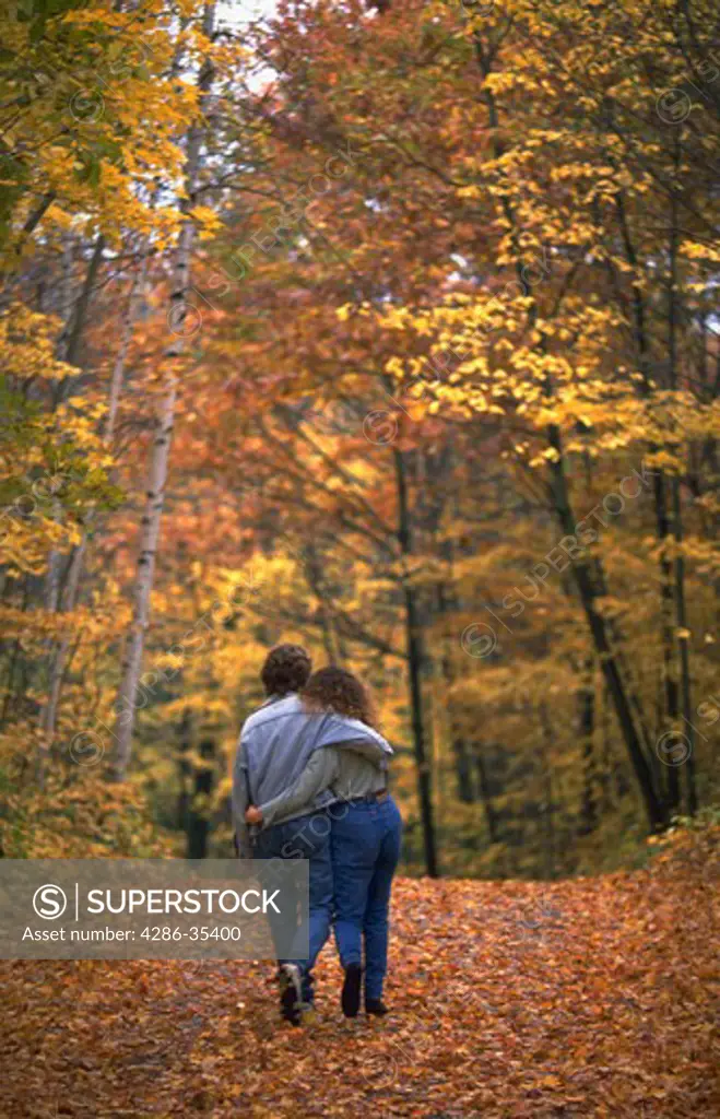 A couple walking with their arms around each other down a tree-lined road with fall foliage.