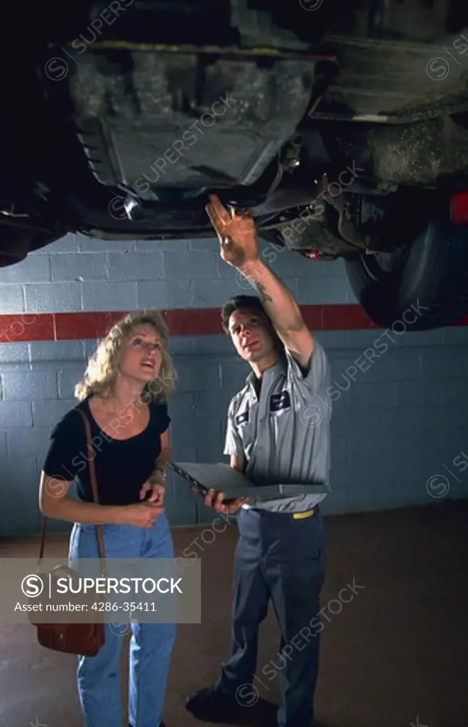 Auto mechanic stands under a car while talking to a female customer about her car.