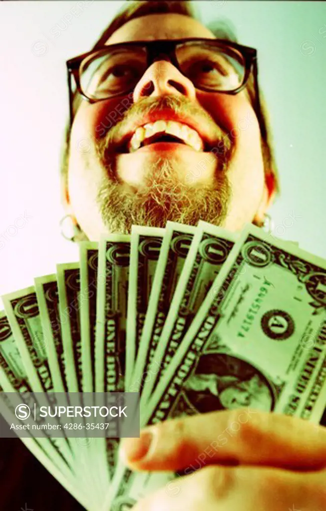 Man wearing glasses with a goatee looks up and smiles while holding a handful of one dollar bills.