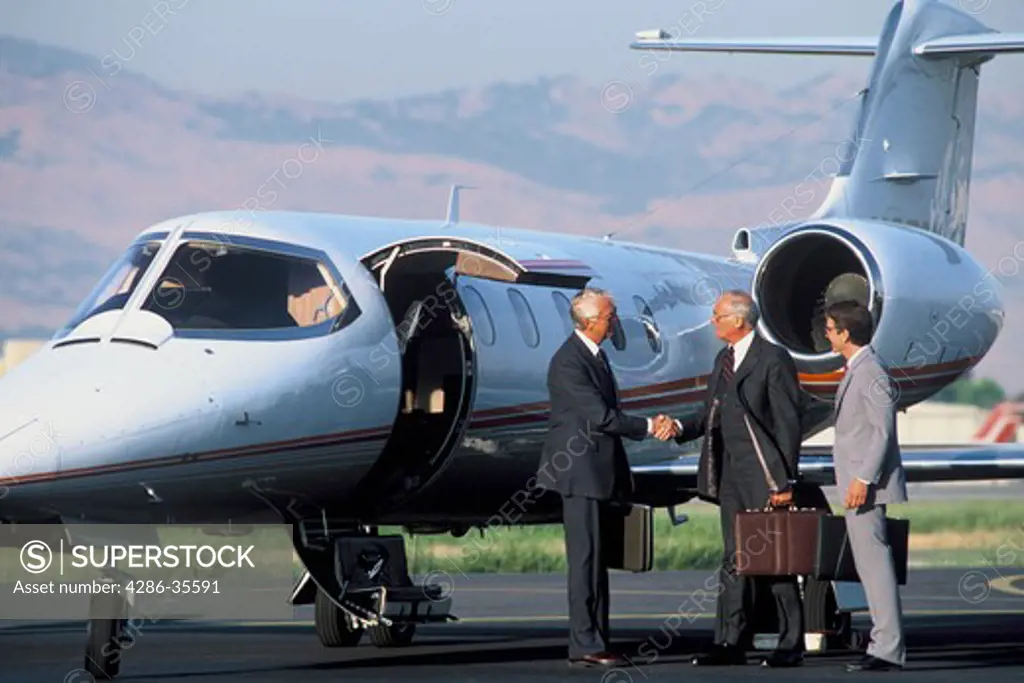 Trio of businessmen standing outside in front of a lear jet with mountains in the background.