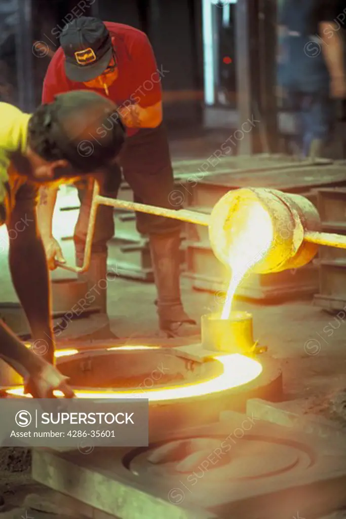 Two steel workers pouring melted steel into large cylinder blocks. 