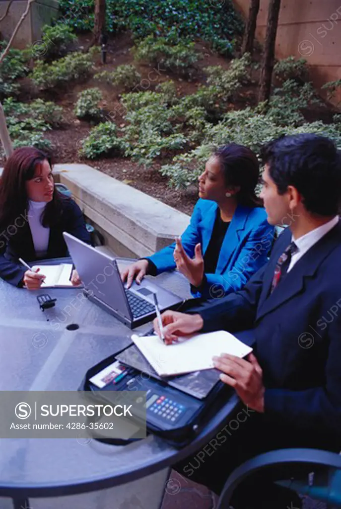 Two businesswomen and a businessman working on their laptops while sitting in an outside table.
