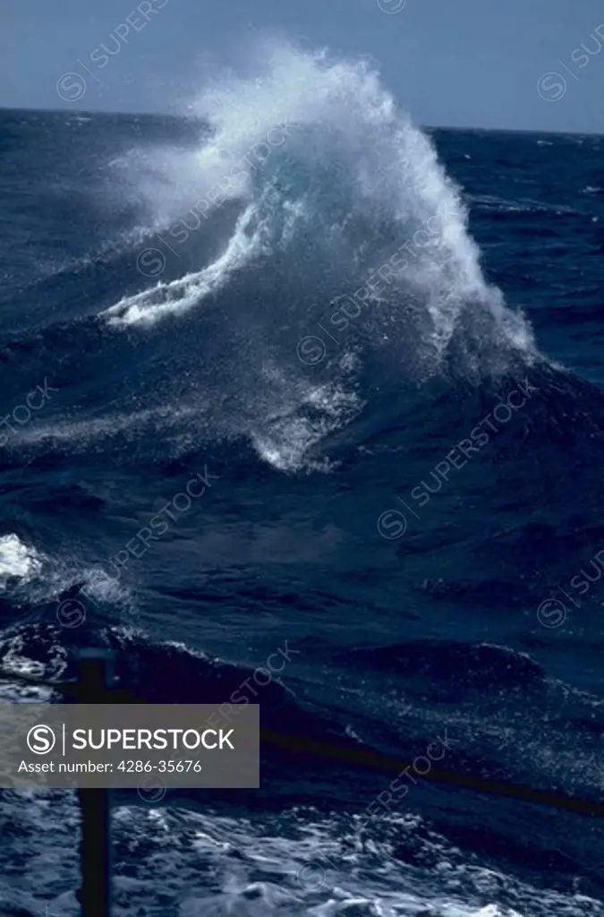 Oceanscape, with dramatic cresting wave, being blown back by wind.  Shows powerful natural force, and heavy seas.  Also shows small detail (a lifeline stanchion) of the ship in lower left foreground from which the shot was made.  (Water Series.  Ocean.  Nature.)