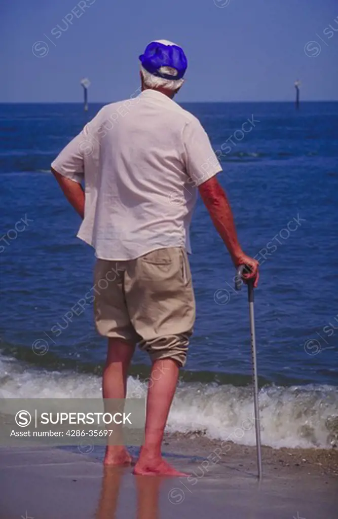 Elderly man, alone.  Hand on a walking cane, with rounded shoulders.   Standing at water's edge, gazing towards the horizon.  Warm weather shot, pants rolled up, barefeet.  Mood of contemplation or personal reflection.  Conveys a certain sense of optimism or vitality with early morning hour and 'getting out and going', plus the youthful ballcap.  Taken in Delaware at Lewes, close to Cape Henlopen.  Note:  John Hartman has an extensive file of water and beach related scenes, illustrating leisure 