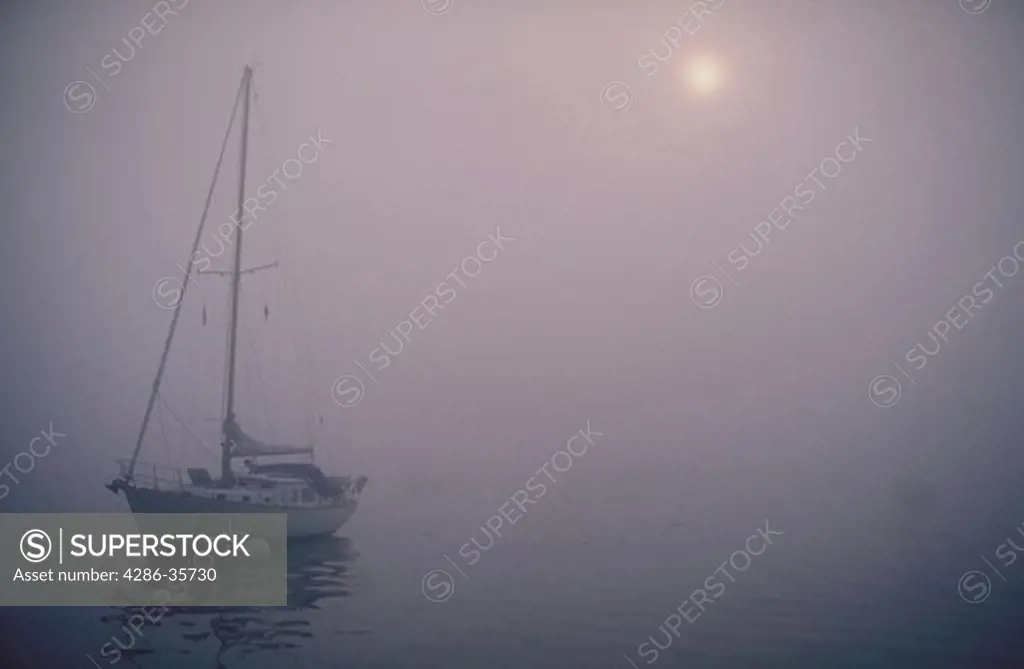 Sailboat, moored, in fog.  Early morning shot with sun beginning to burn through the fog.  Taken in Boothbay Harbor, Maine.   Other heavy fog photos available. 