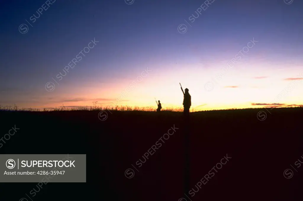 Silhouette of two pheasant hunters in a field at sunrise.