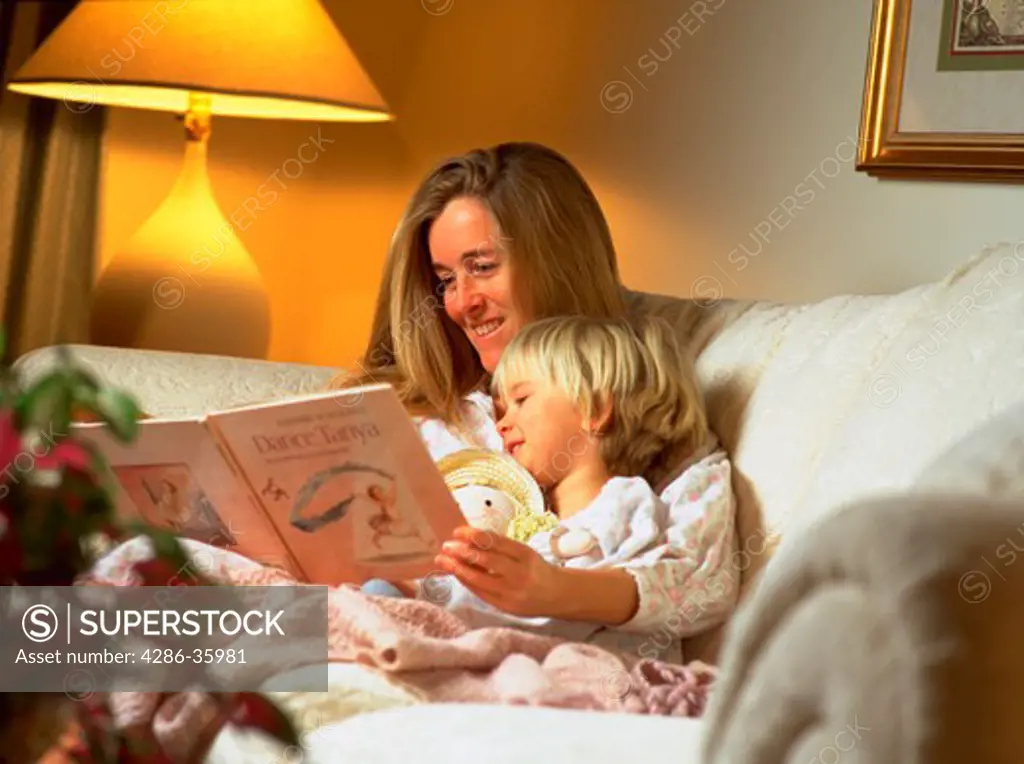 Mother reading book to young daughter while sitting on the couch.