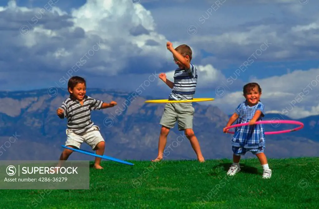 Three children playing with hula hoops on a grassy knoll.