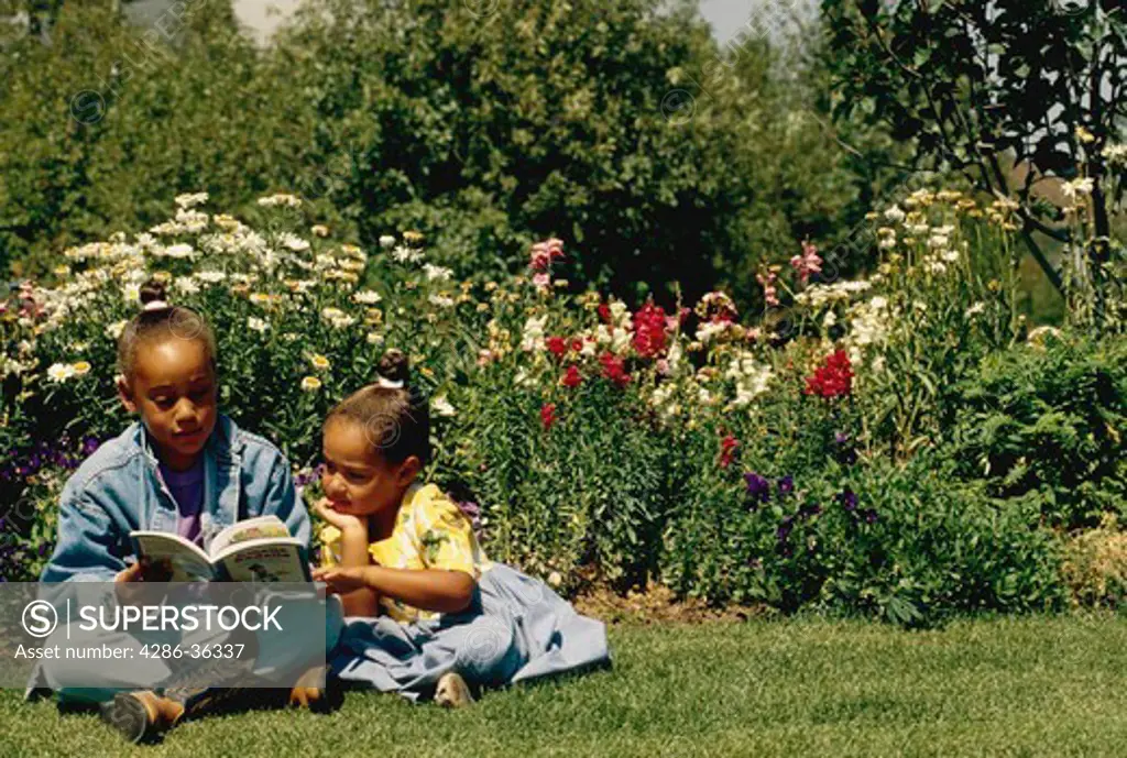 Two young, African-American girls reading a book outside next to a flower bed. 