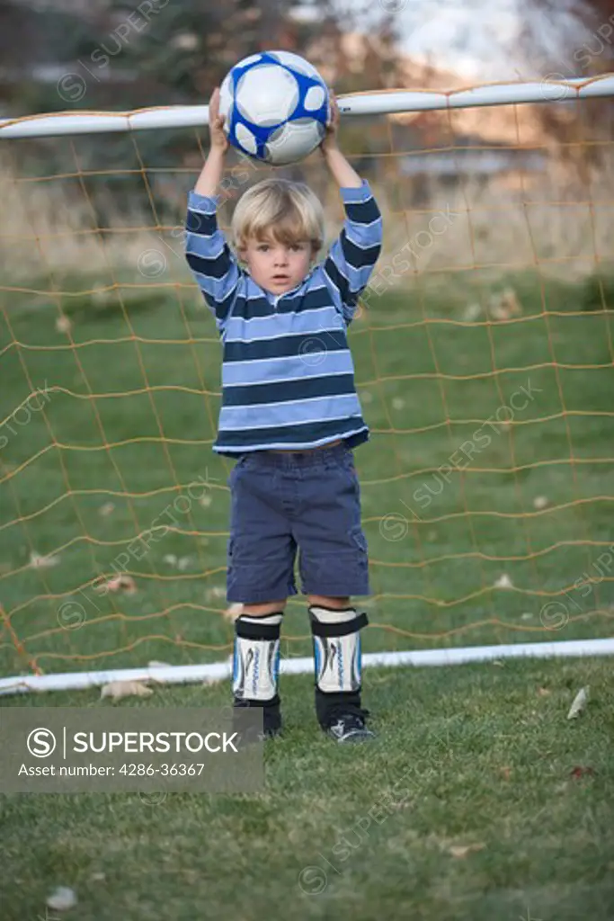 Little blond boy holding a soccer ball over his head standing in front of the goal. 