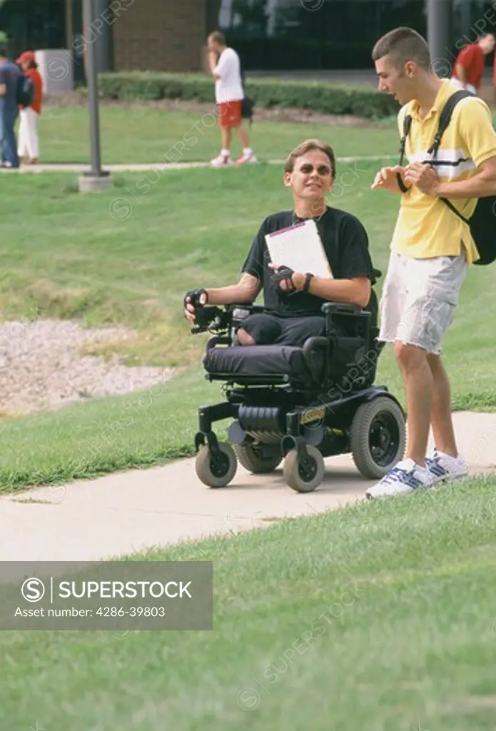 Paraplegic on an electric wheelchair talking with another student outdoors. 