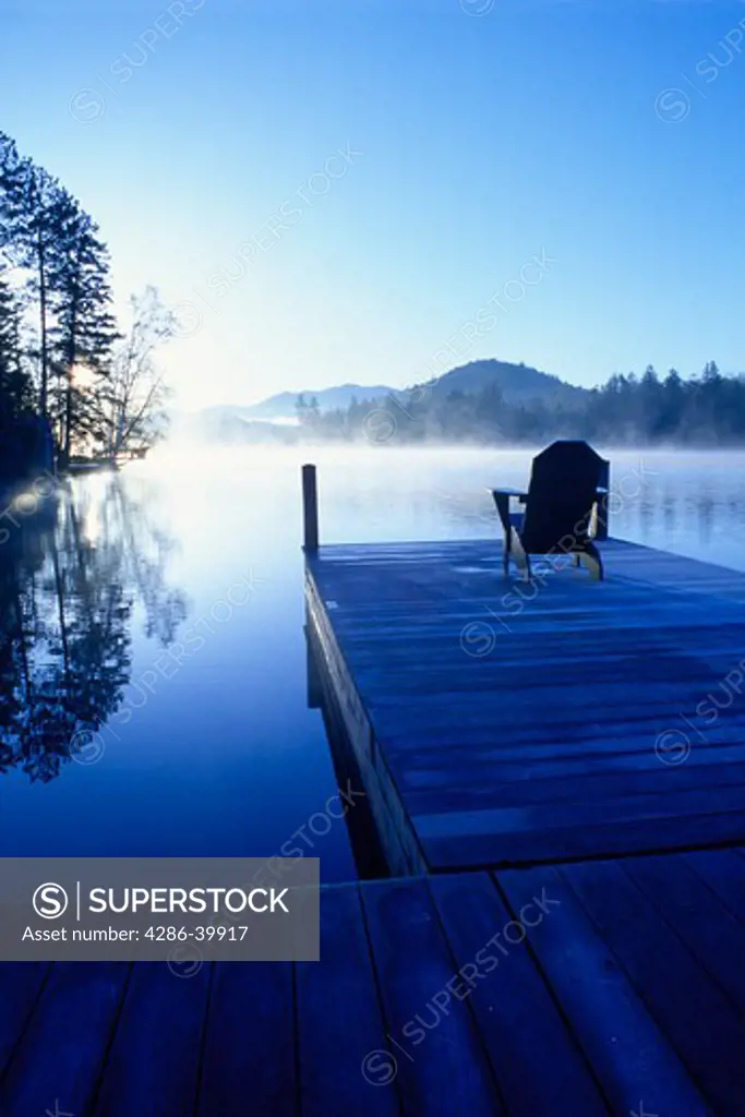 Empty deck chair sits on dock by misty Mirror Lake, Lake Placid, NY overlooking sunrise, trees and hills. 