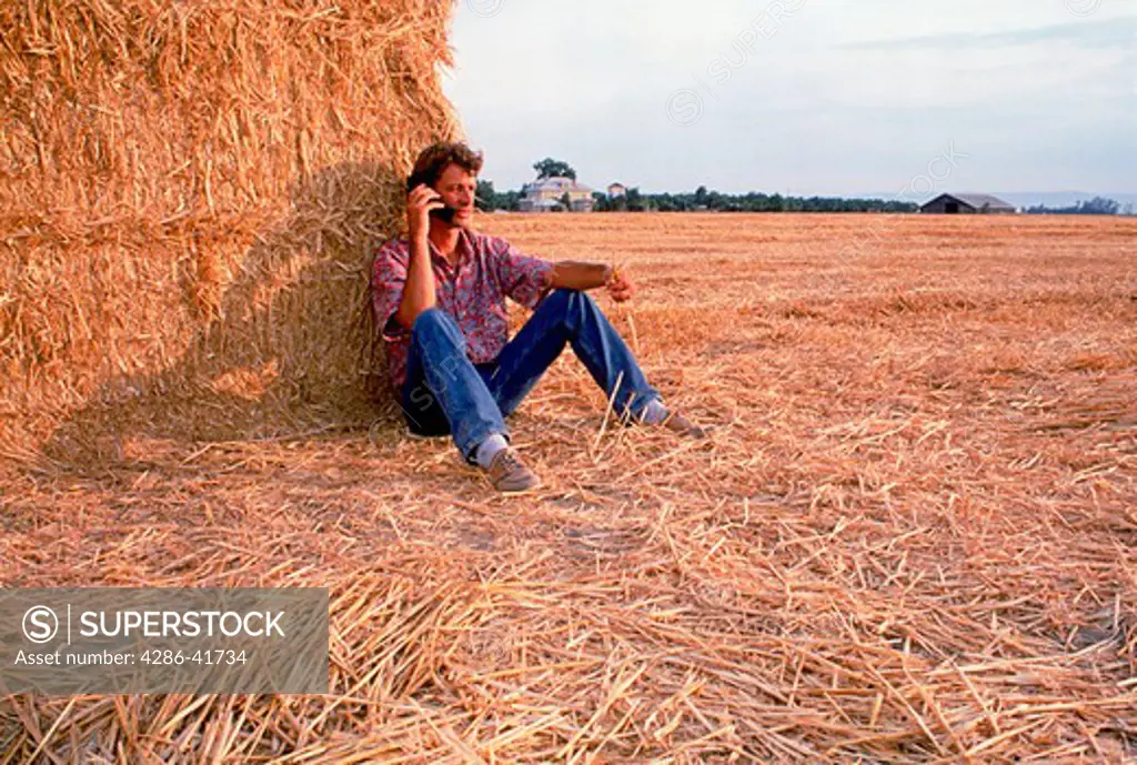 young farmer with cellular flip-phone sitting on straw bales