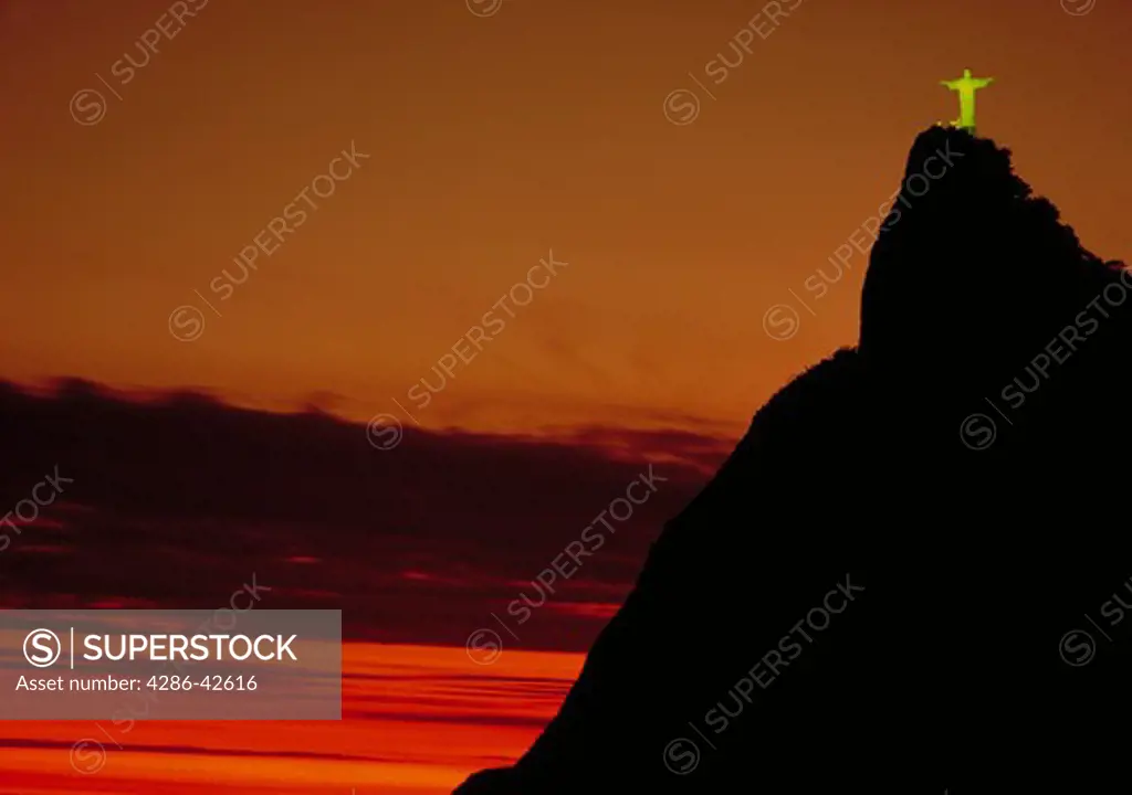 The statue of Christ the Redeemer (Corcovado) standing high atop a hill at sunset against an orange sky in Rio de Janeiro, Brazil.