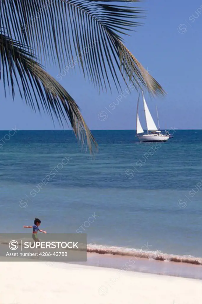 Child walking along white sandy beach, with sailboat in background.