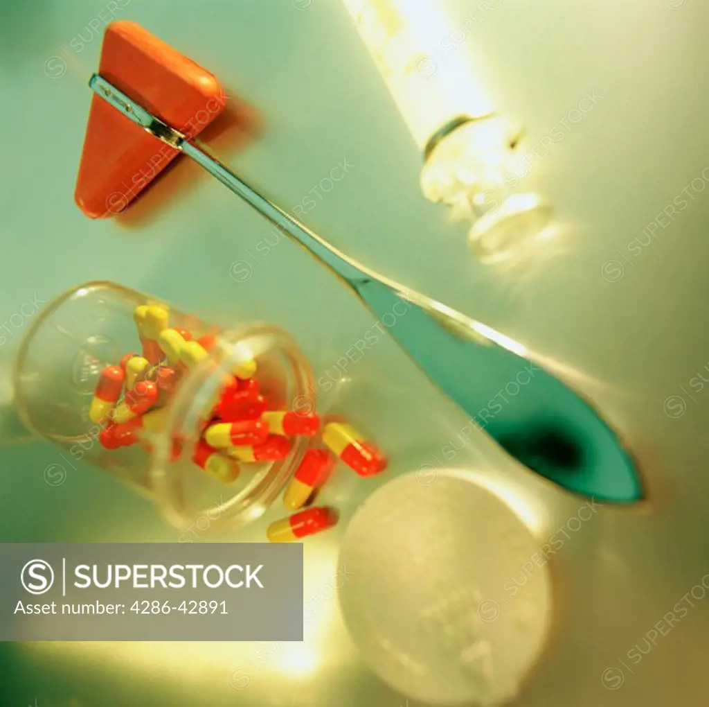 Still life of a doctor's rubber hammer, red and yellow capsules spilling out of an opened vial, and a hypodermic syringe.