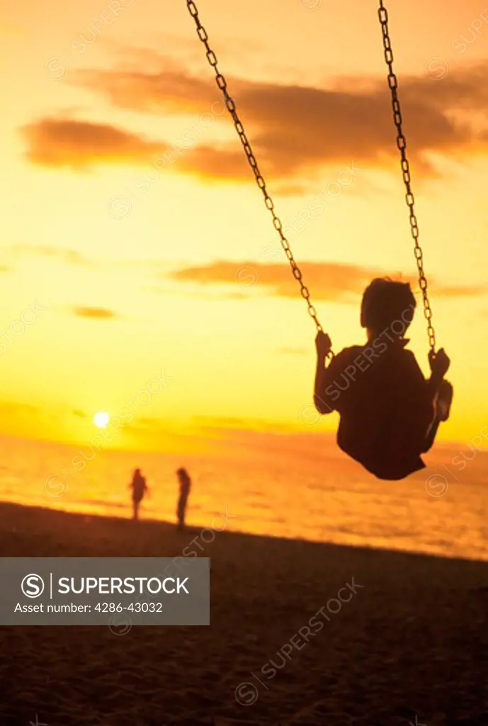 Silhouette of a child on a swing at the beach as  sun sets over the ocean with a couple in the distance.