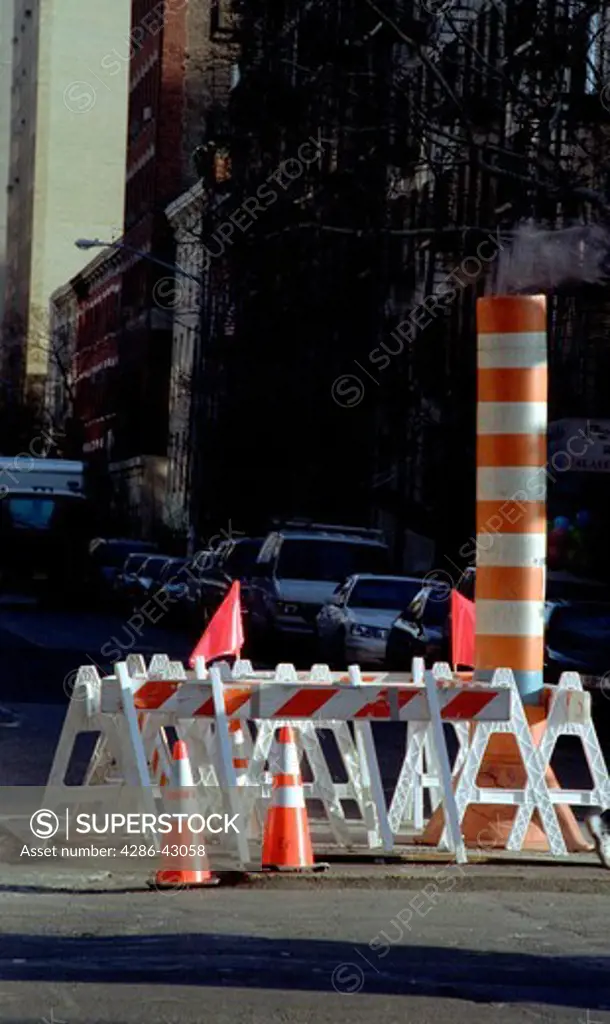 Cautionary sawhorses and bollards surround a temporary steam vent in the middle of a city street during road repairs.