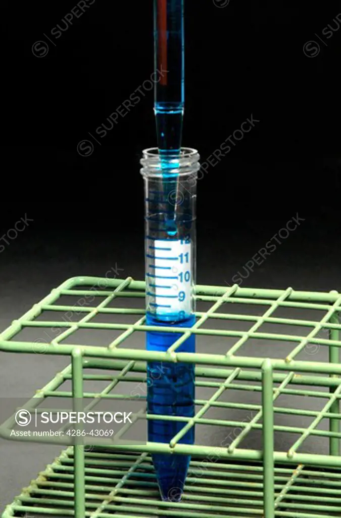 A pipette is used to pour a blue liquid into a test tube which stands alone in a wireframe holder.