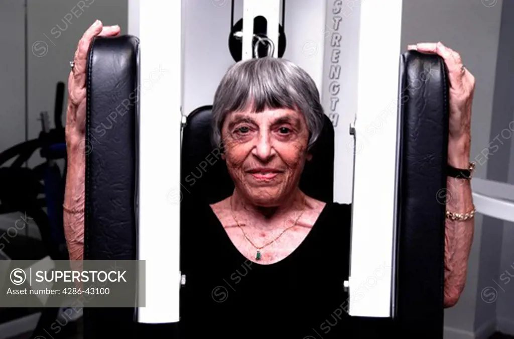 A smiling senior woman in black leotard exercising her arms on a PecDec machine.