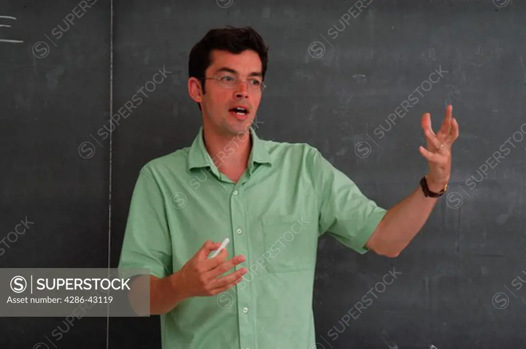 Young male teacher standing in front of the chalkboard lecturing to an unseen audience. 