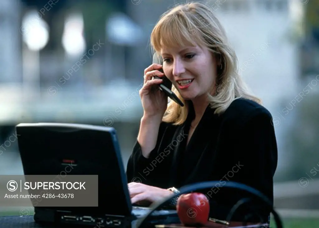 Woman talking on cellphone outdoors with laptop computer