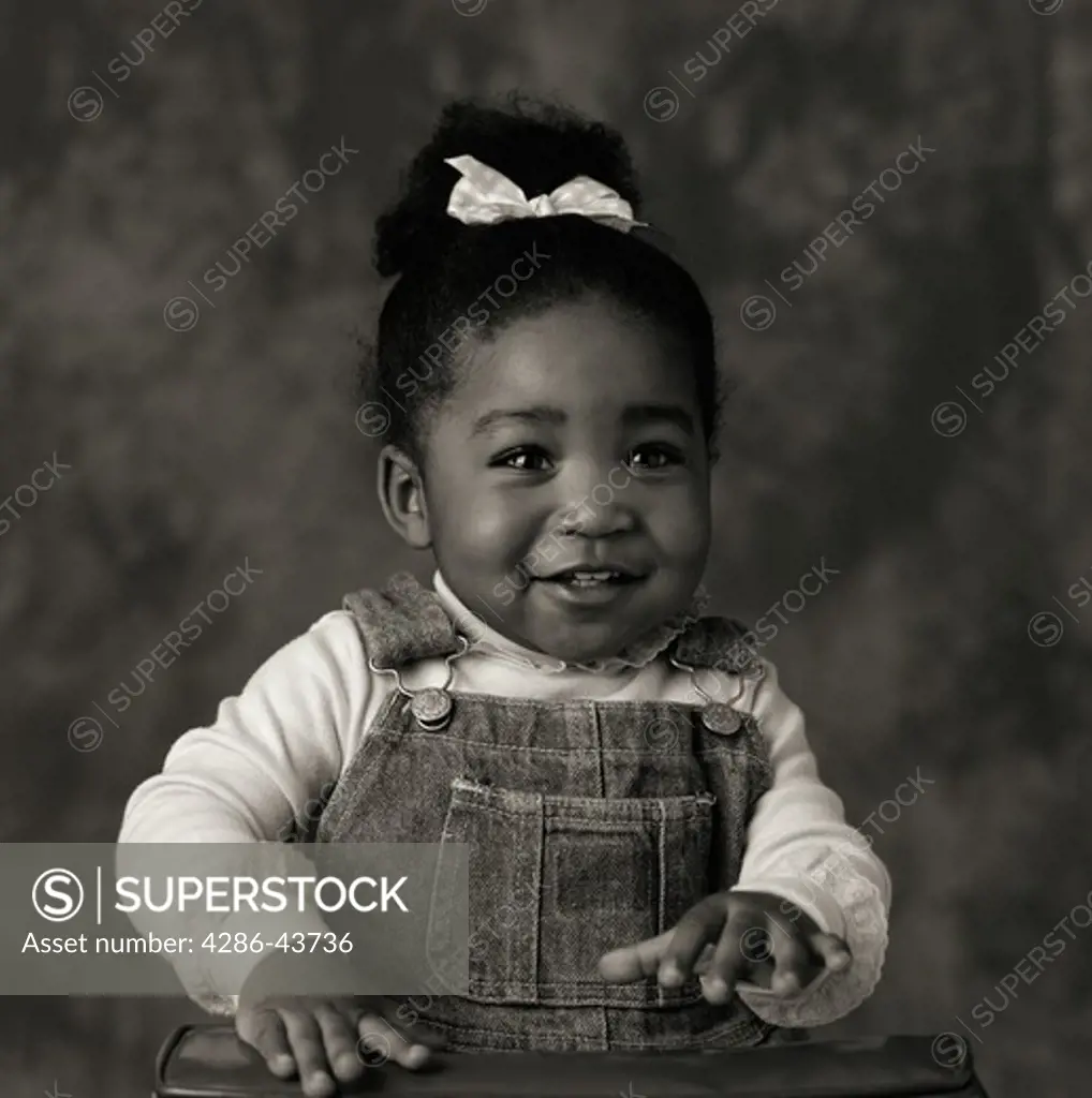 Black and white portrait of a young African American girl in overalls and a bow in her hair.
