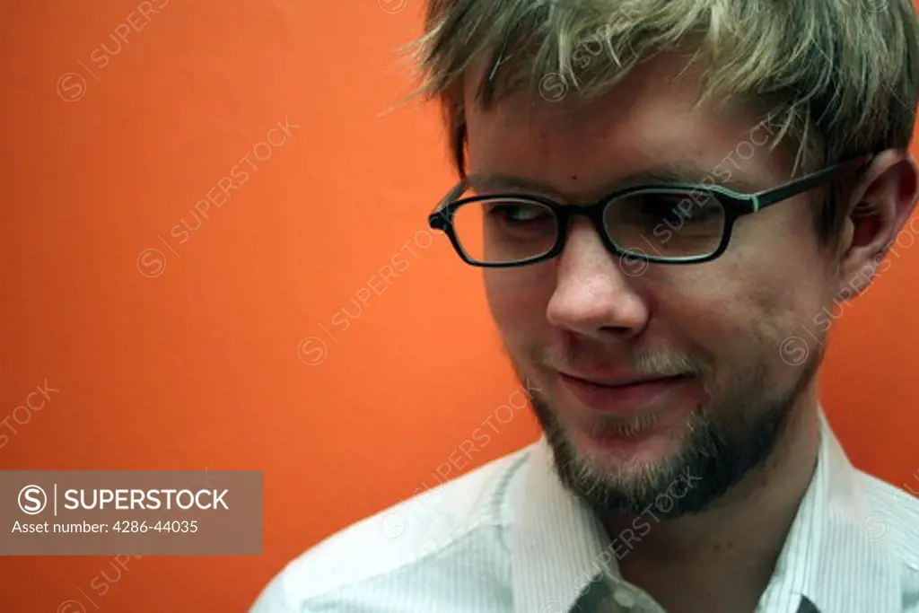 Young blonde man with plastic glasses frames smiles while standing against an orange wall