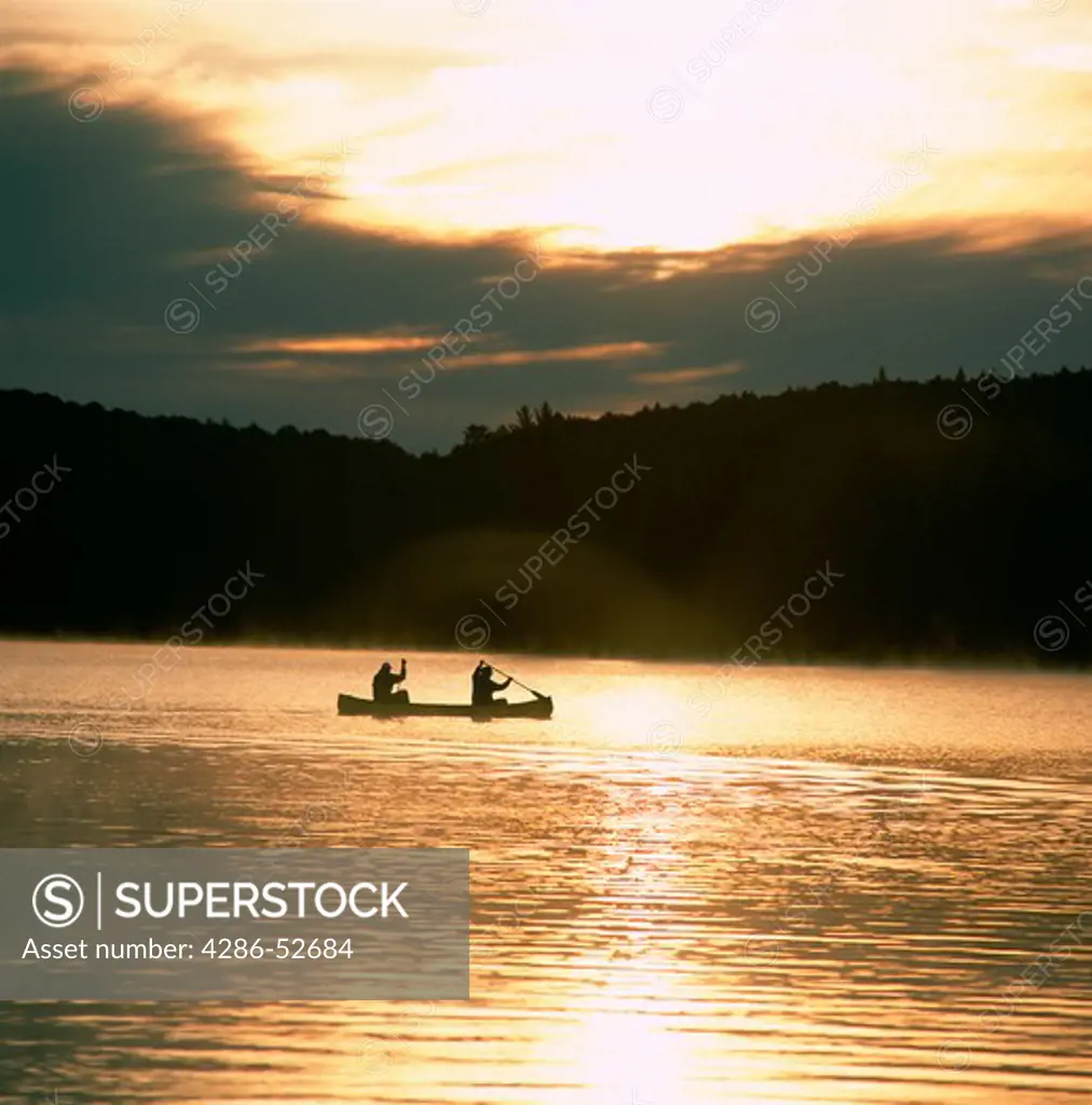Two canoeists paddling on a misty lake in the dawn's early light in Algonquin Park, Ontario, Canada.