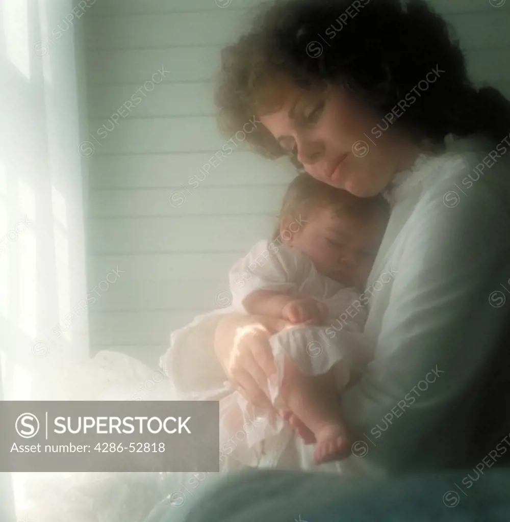 Mother holding her sleeping, infant daughter as she sits by the window. 