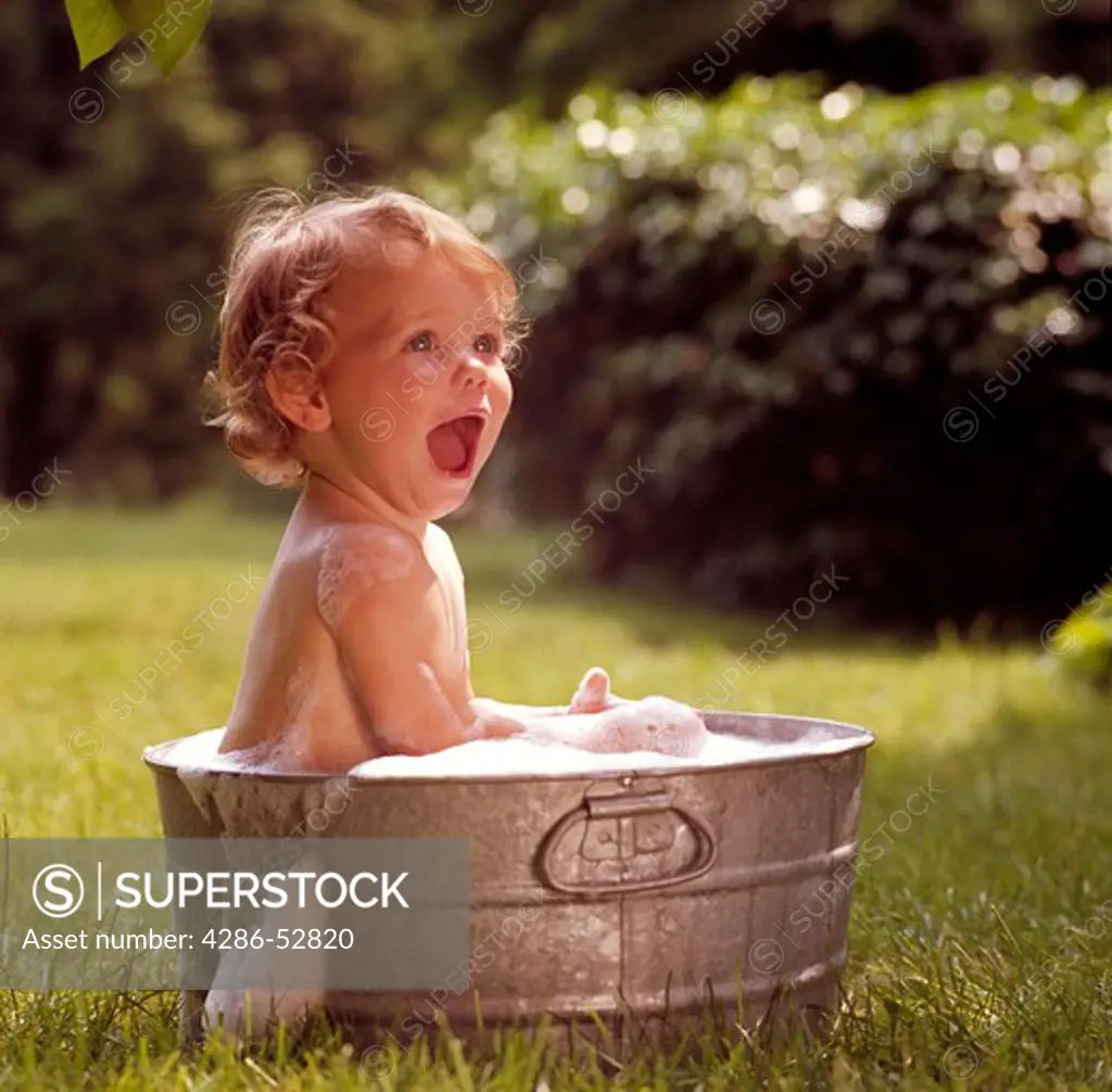 Toddler sitting in an aluminum tub outside taking a bath as the soap suds overflow. 