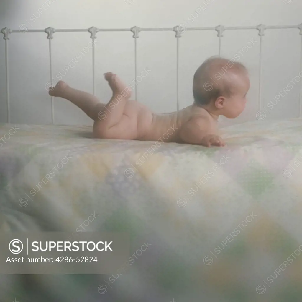 A  naked baby  lying on its tummy on a bed while kicking its legs in the air.