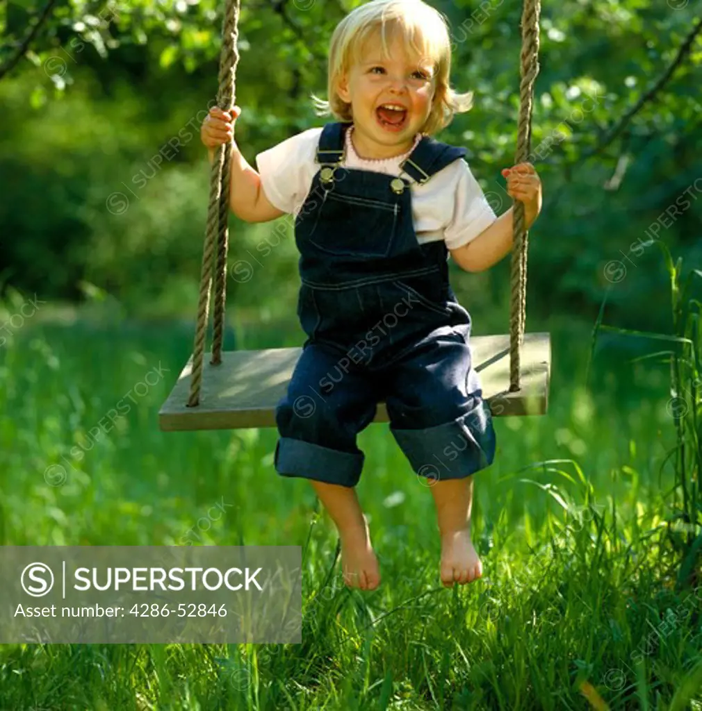 Little girl wearing overalls and swinging on a wooden swing with her feet dangling over grass blades. 