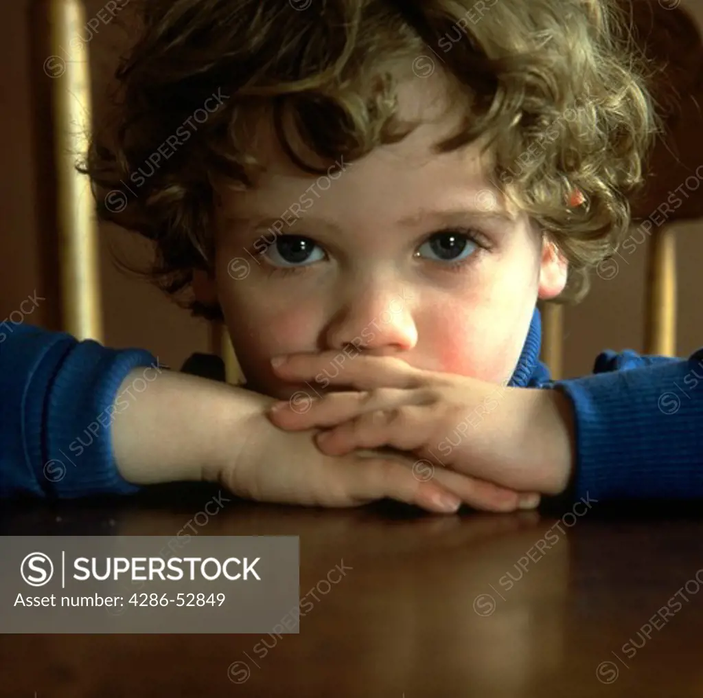 Young boy with arms crossed on the table and resting his head on his arms looking directly at the camera.