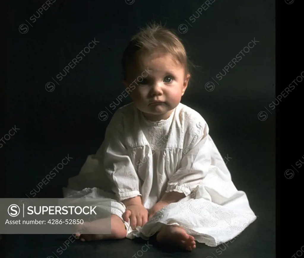 Infant wearing a white gown sitting on the floor in the dark. 