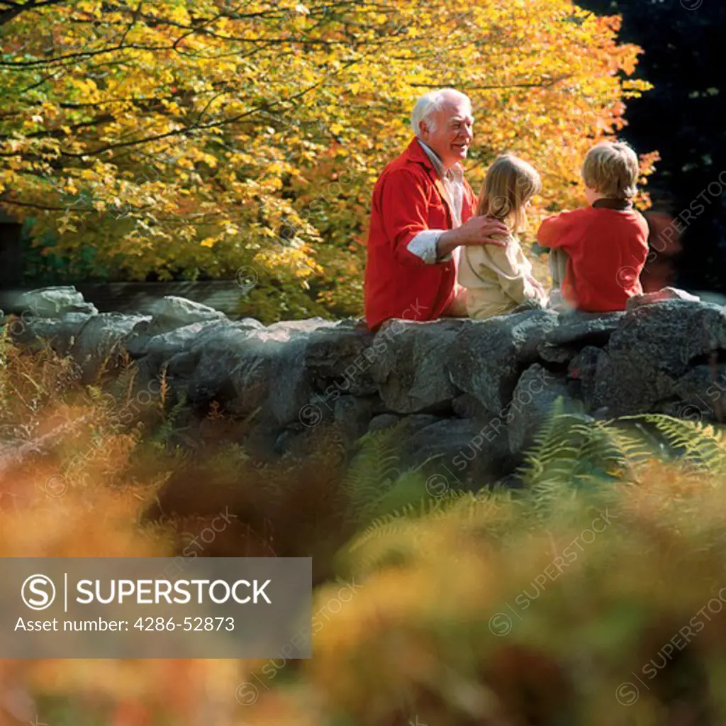 A grandfather sitting amongst the fall foliage on a rock wall with his grandson and granddaughter.