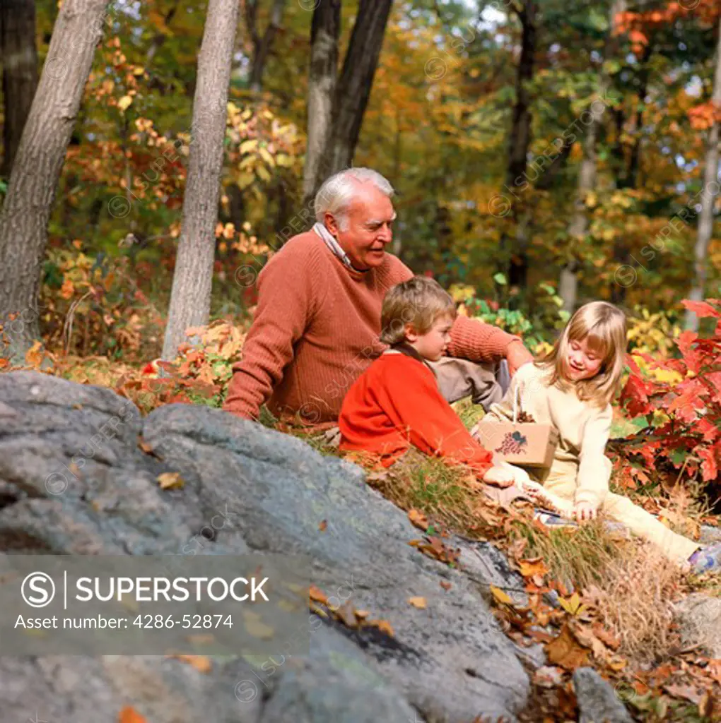 A grandfather witting amongst the fall foliage with his grandson and granddaughter.