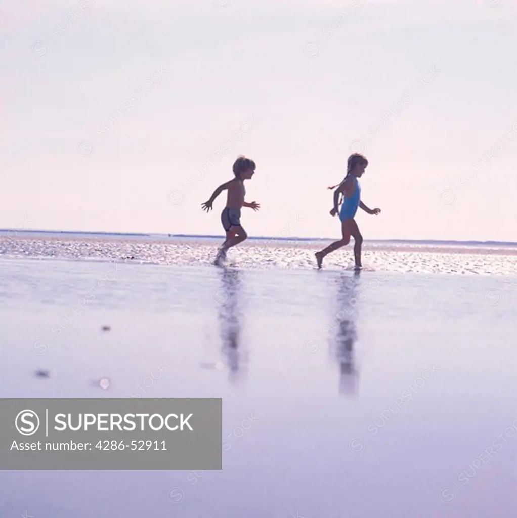 A young boy and young girl running through shallow water at the beach.