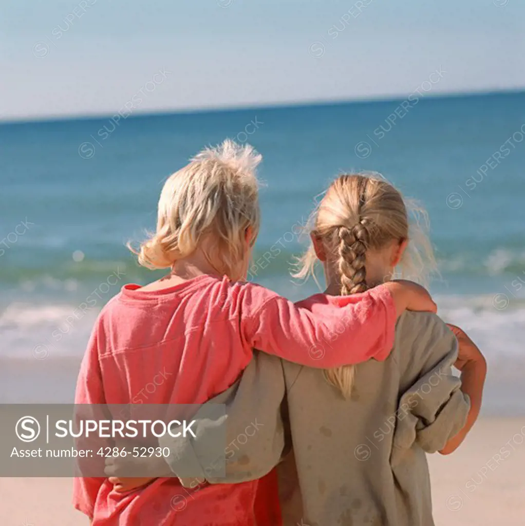 View from behind as a young boy and a young girl stand arm-in-arm on the sandy shore.