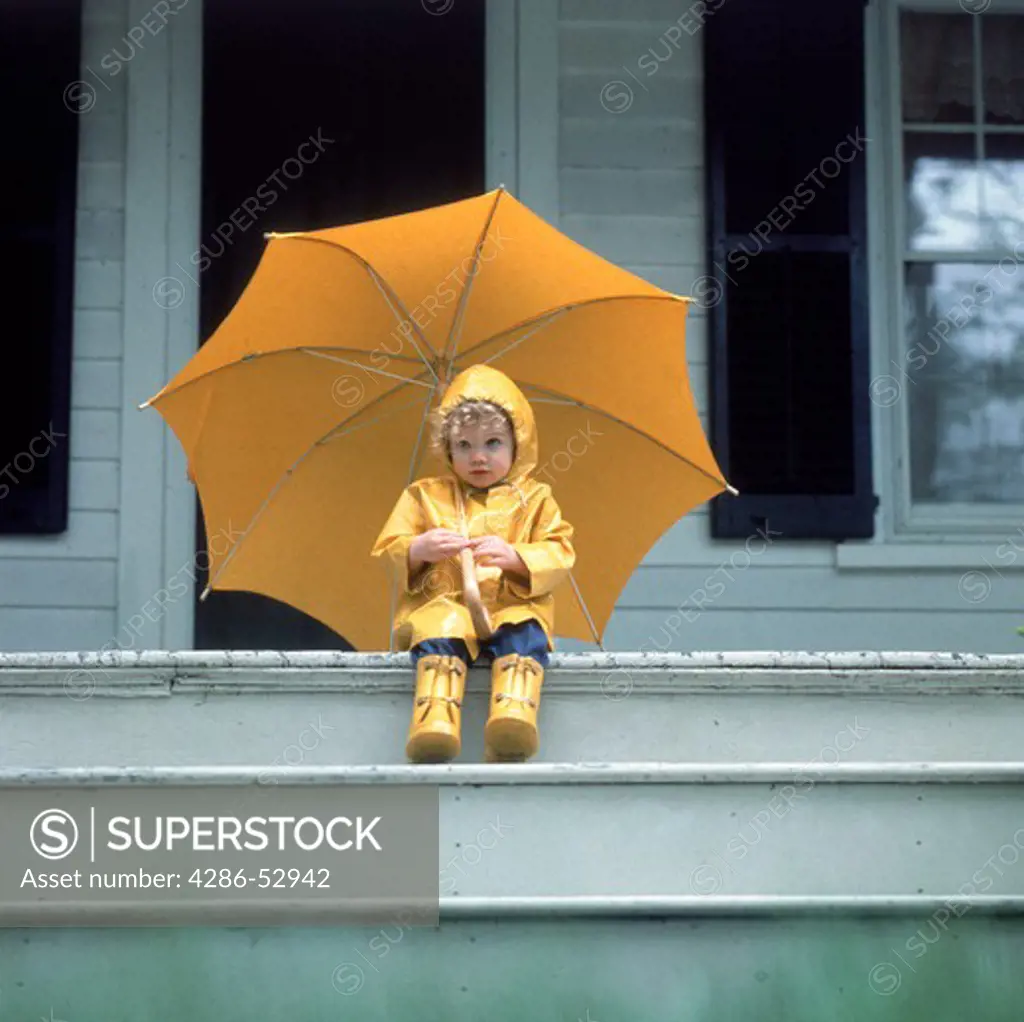Portrait of a young child wearing a yellow rain coat and rain boots holding an umbrella sitting on the front porch of a house.
