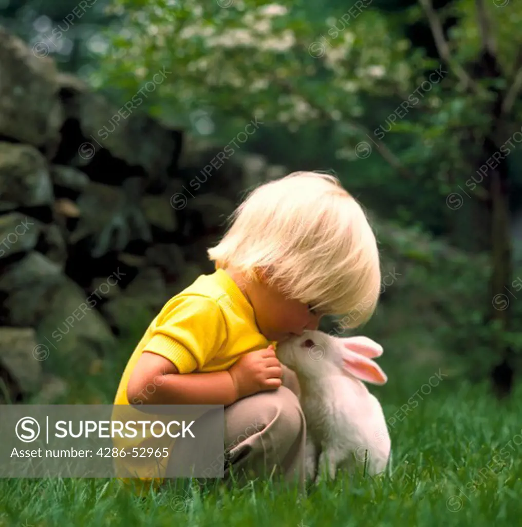 Little, blonde boy kneeling down over and kissing a white bunny rabbit on its nose while in the grass.