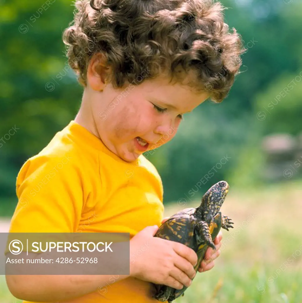 Curly-haired boy wearing a yellow t-shirt with dirt on his arms and face holding an Eastern Box turtle.