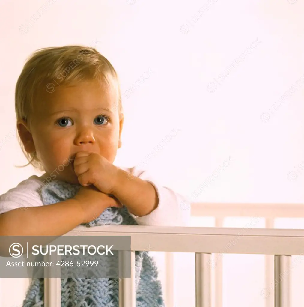 Baby holding a blue blanket while standing up in the crib with a look of concern on the face. 