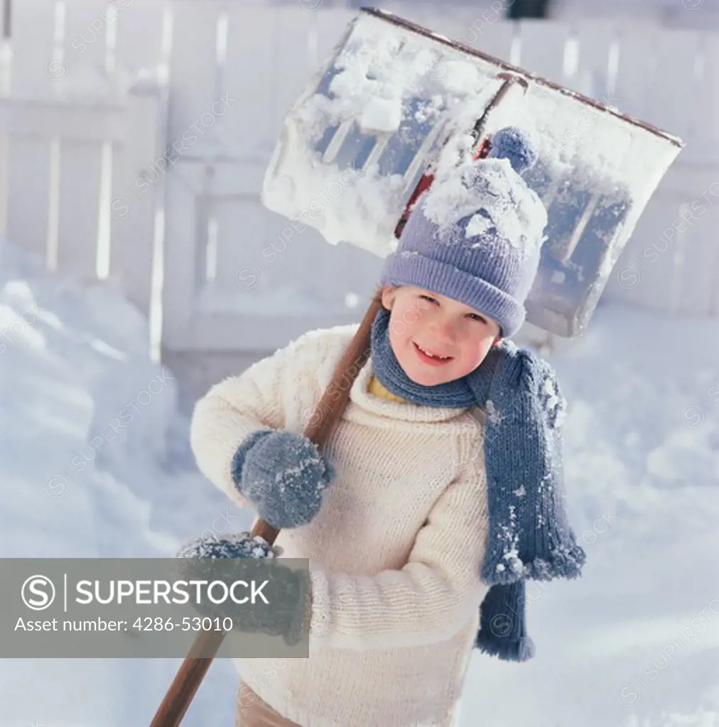 Smiling child grips a snowshovel in his blue knit mittens as he stands outside on a sunny snowy day.